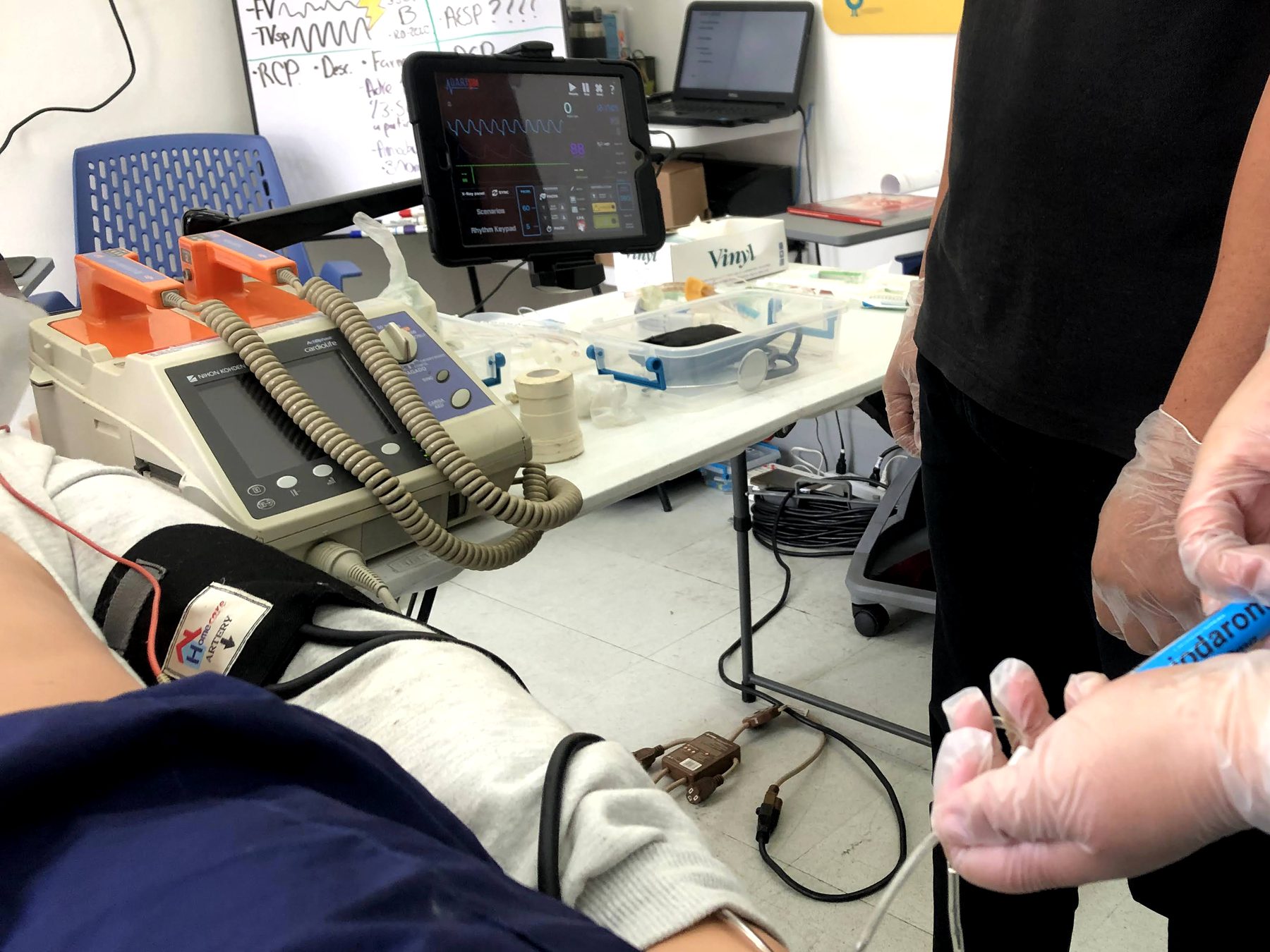 Práctica de ACLS: monitor desfibrilador, simulador de ritmos en una tableta y manos con guantes preparando un medicamento.