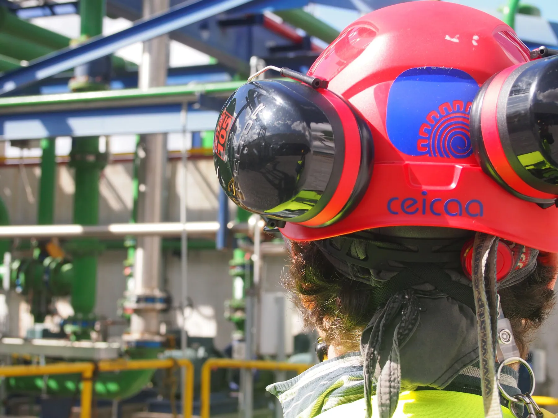 Casco rojo de rescate con el logo de CEICAA y orejeras, visto por detrás, durante una práctica en una planta industrial.