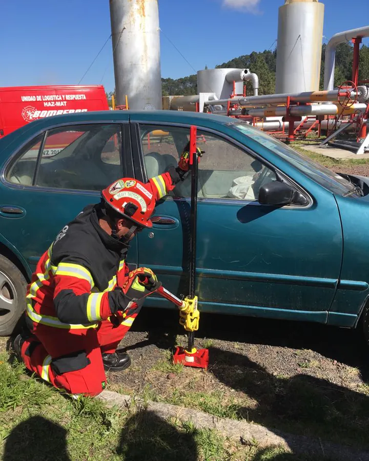 Bombero con equipo estructural opera un gato Hi-Lift junto a un auto en una planta industrial.