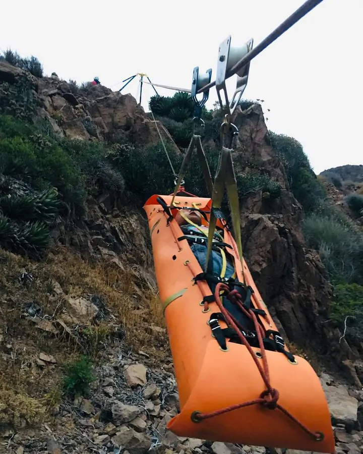 Camilla naranja tipo canastilla colgada de una tirolesa sobre una ladera rocosa.