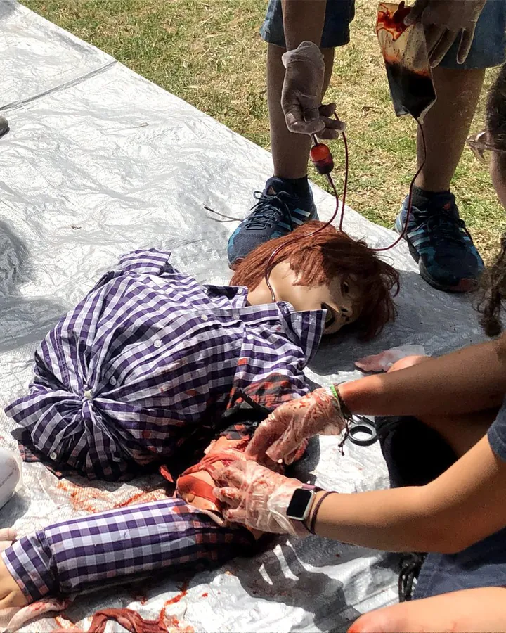 Manos con guantes atienden una hemorragia simulada en una víctima de práctica sobre una lona.