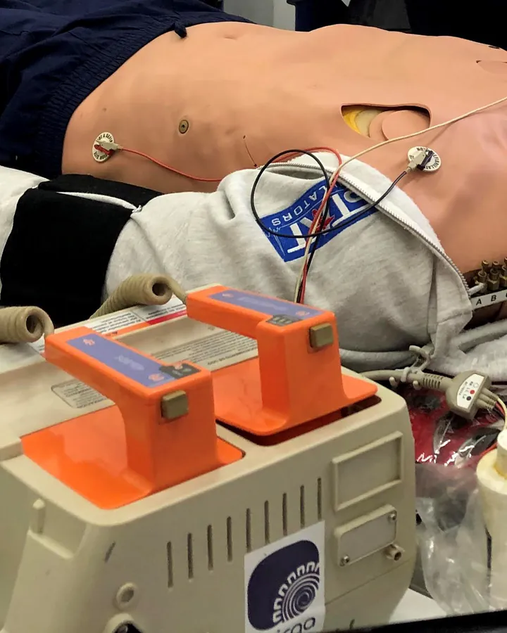Manos sobre un maniquí avanzado en camilla, con monitor desfibrilador, electrodos y bolsa-válvula.