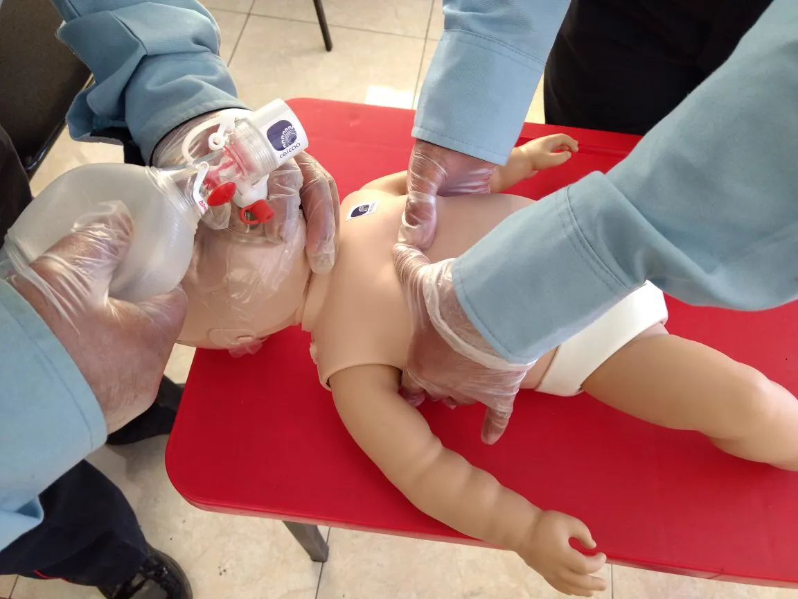 RCP en un maniquí de lactante: una persona ventila con bolsa-válvula pediátrica y otra da compresiones con dos pulgares.