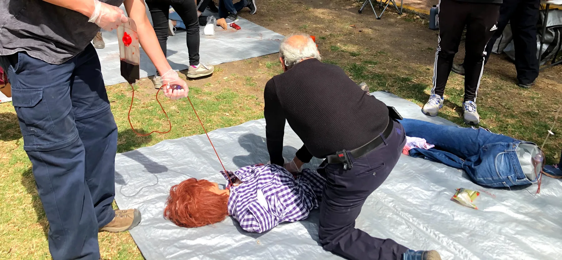 Práctica al aire libre: un participante presiona la herida de un maniquí sobre una lona mientras otro sostiene la bolsa de sangre simulada.