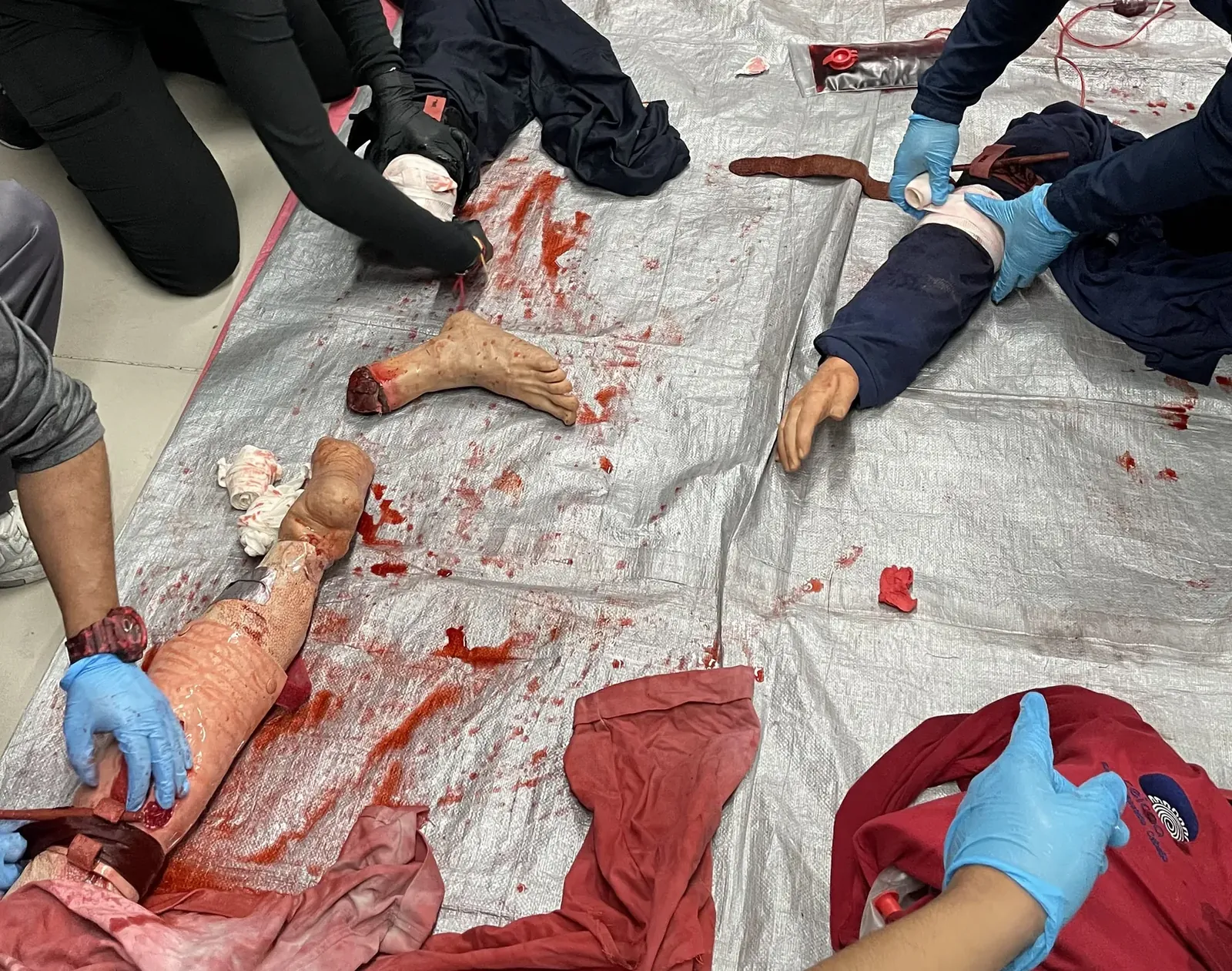 Estación de control de hemorragias del PHTLS: manos con guantes trabajan sobre simuladores de extremidades con sangrado, sobre una lona.