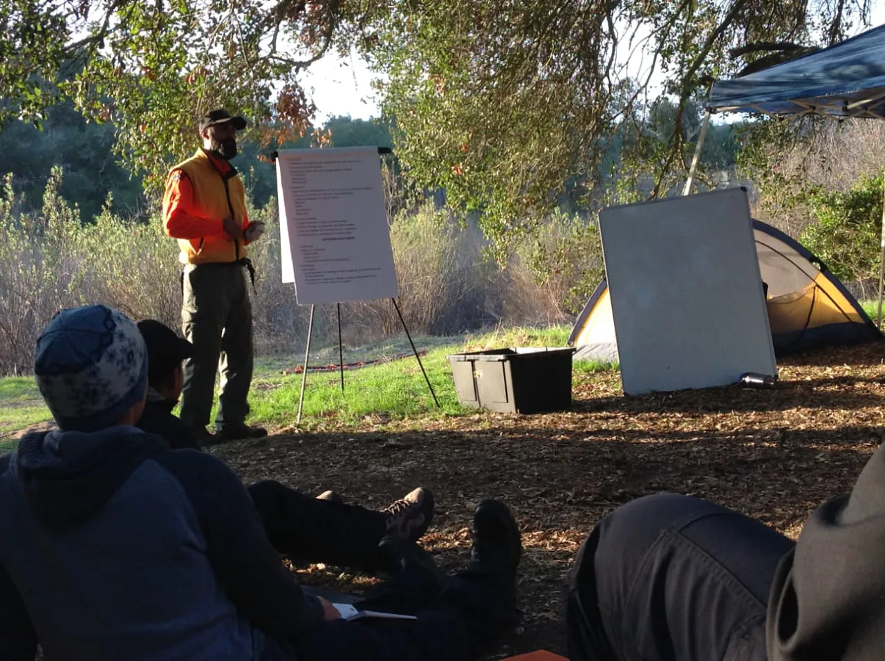 Clase al aire libre: el instructor explica junto a un rotafolio y el grupo escucha sentado, de espaldas.