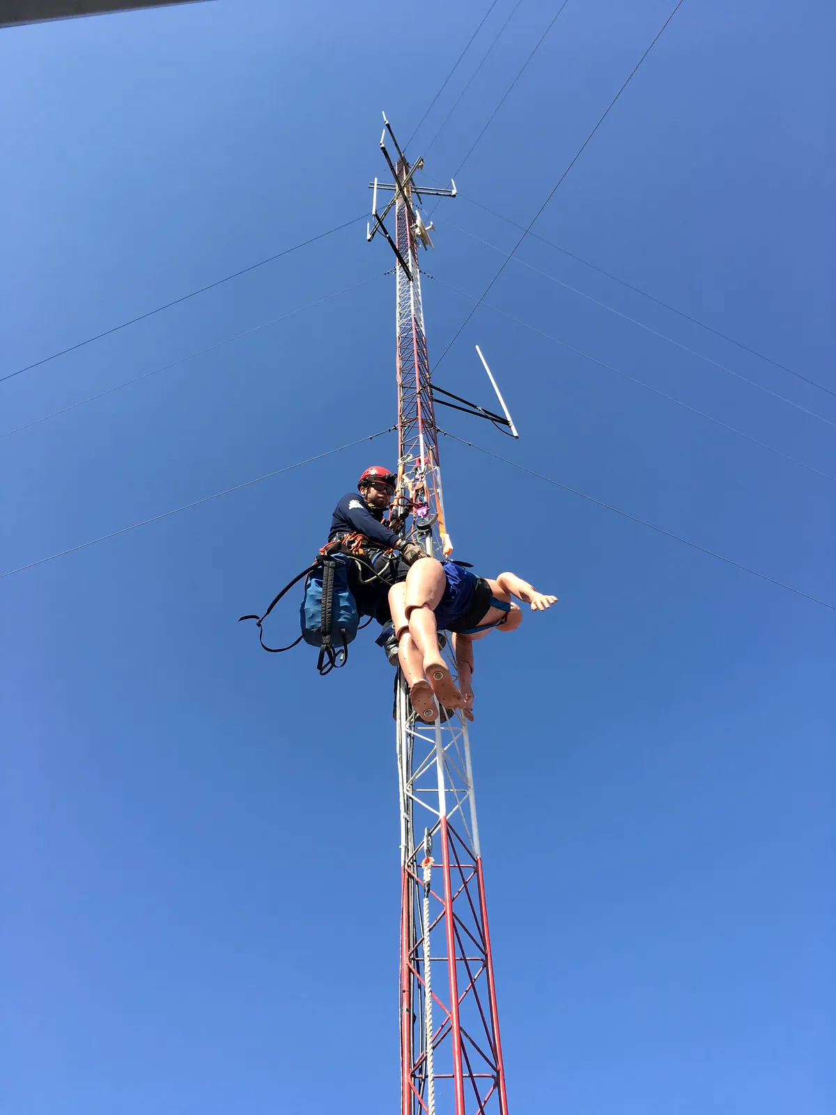 Rescatista suspendido en una torre de telecomunicaciones baja a un maniquí, contra el cielo azul: práctica de rescate en altura.