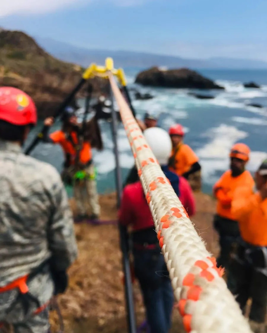 Cuerda tensada en primer plano hacia el mar, con rescatistas de naranja y casco rojo, desenfocados, en la costa.