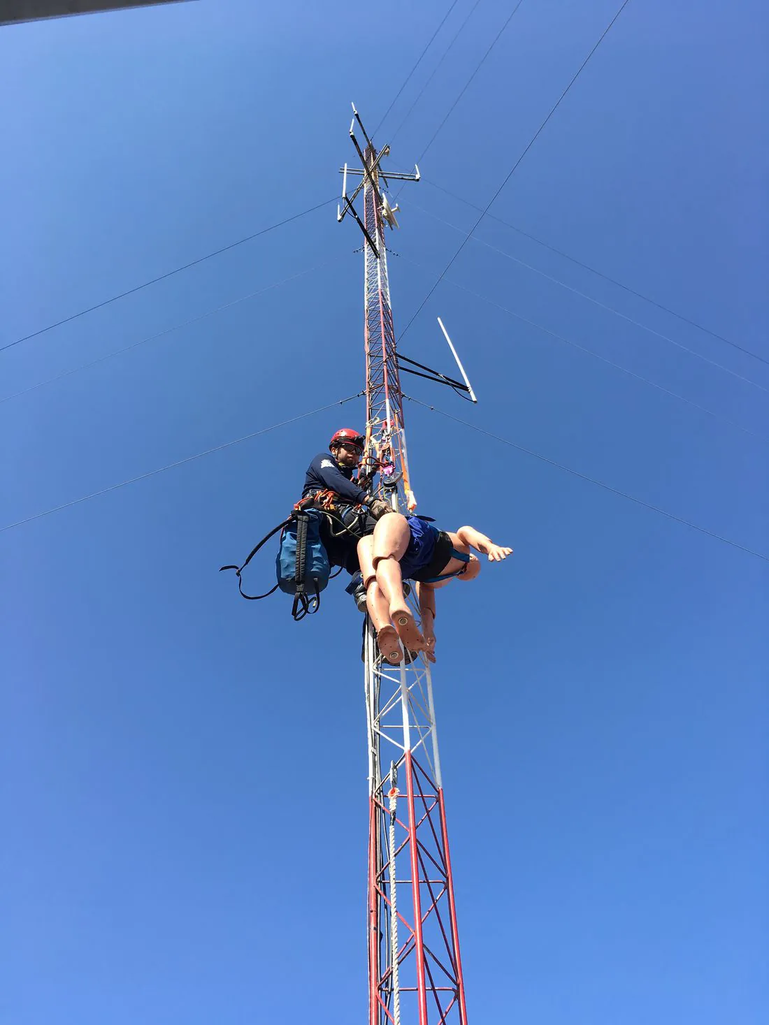 Rescatista suspendido en una torre de telecomunicaciones con un maniquí, contra el cielo azul.