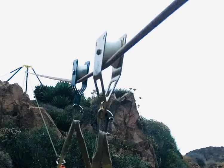 Poleas sobre una línea tensada y un trípode de anclaje en lo alto de una ladera rocosa.