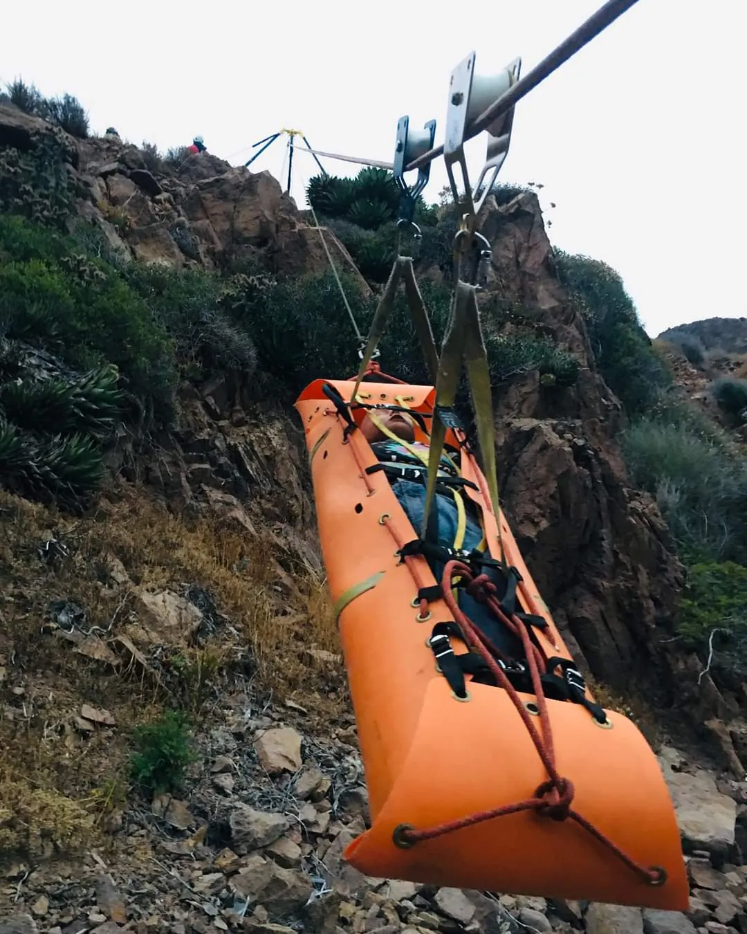 Camilla naranja de rescate colgada de una tirolesa sobre una ladera rocosa, con el trípode y el equipo en lo alto.