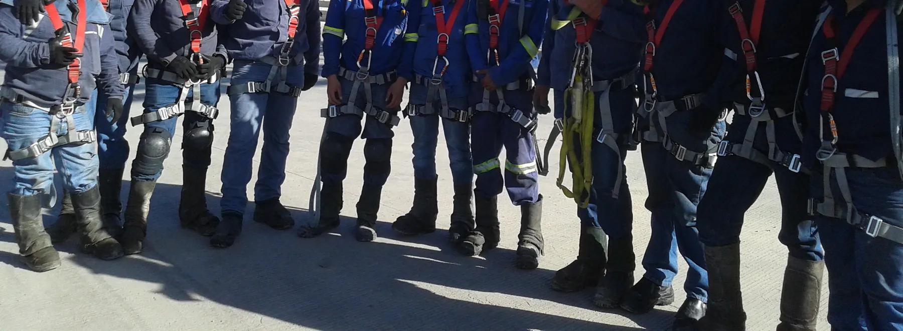 Fila de trabajadores con arnés de cuerpo completo, guantes y botas en el patio de una planta industrial.