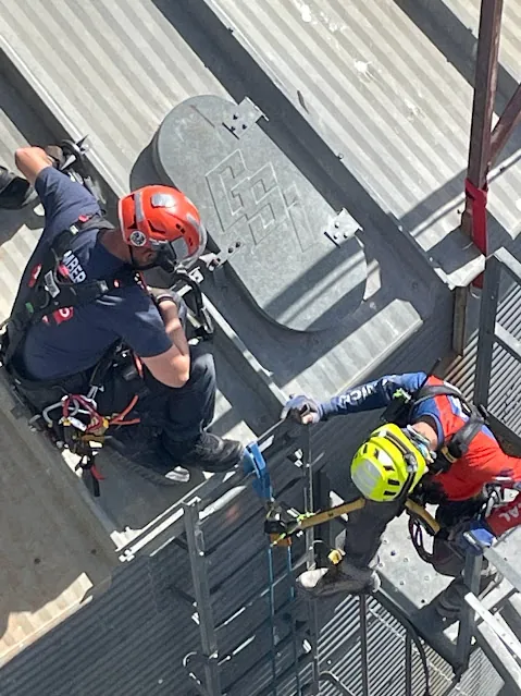 Vista desde arriba: dos rescatistas con casco suben por la escalera de un silo junto a la escotilla.