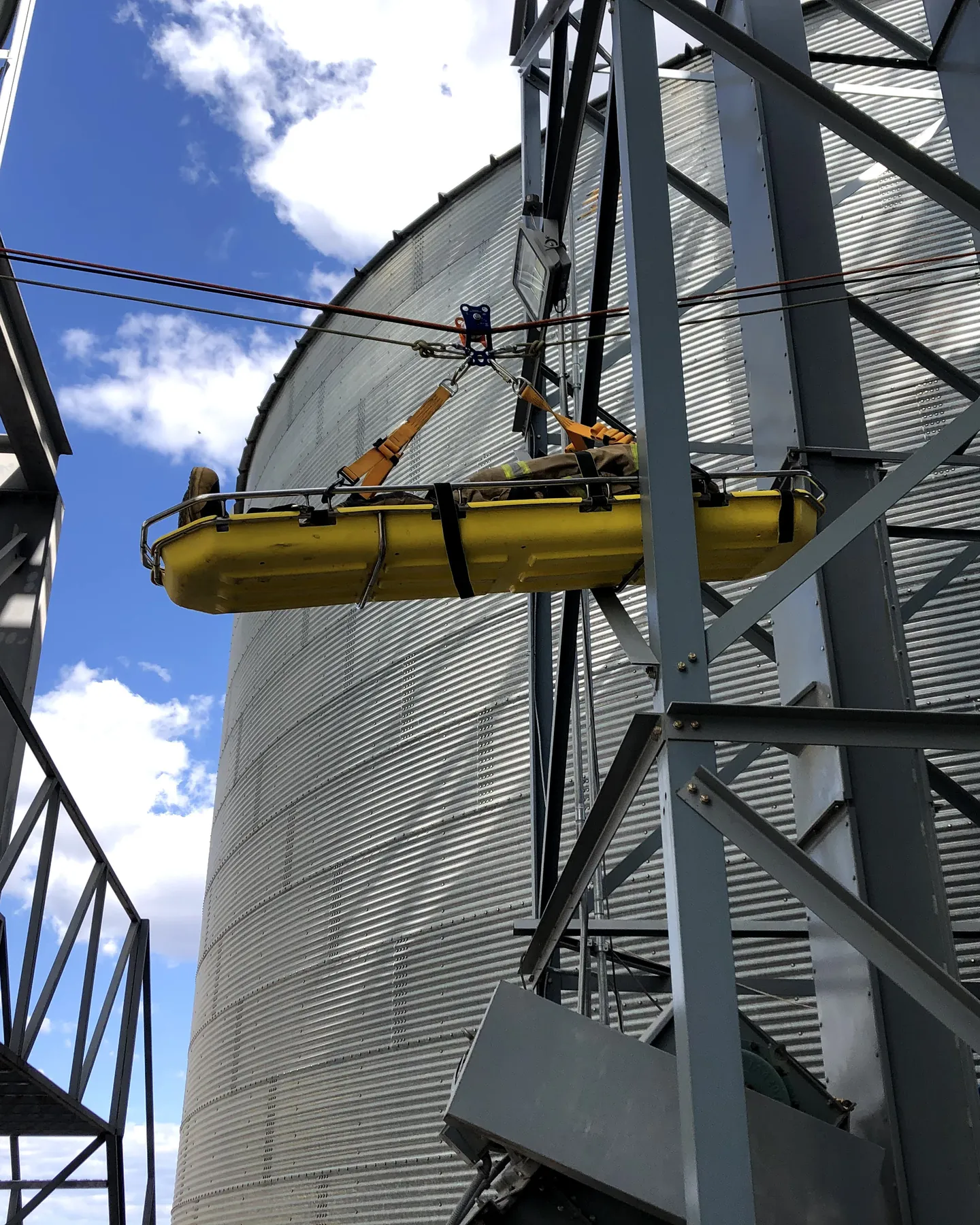 Camilla amarilla con una víctima simulada izada entre dos silos de granos y su estructura metálica, con el cielo azul.