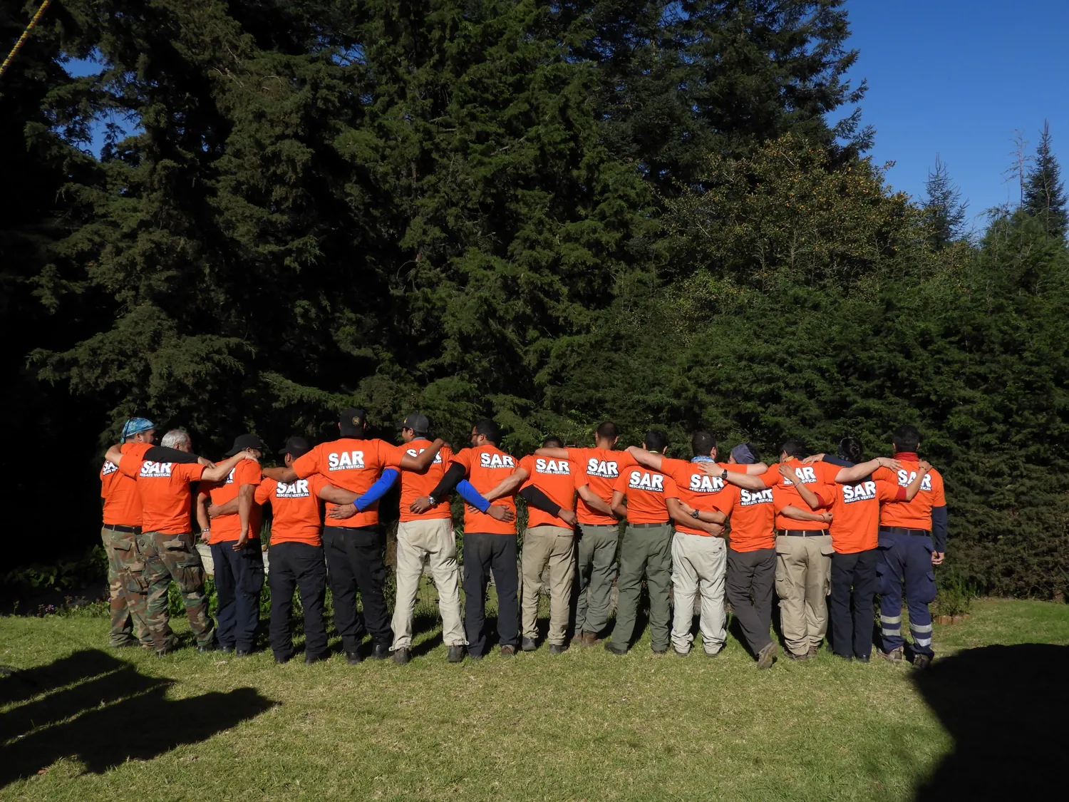 Grupo de unas quince personas de espaldas, abrazadas, con playeras naranjas que dicen «SAR», en un claro del bosque.