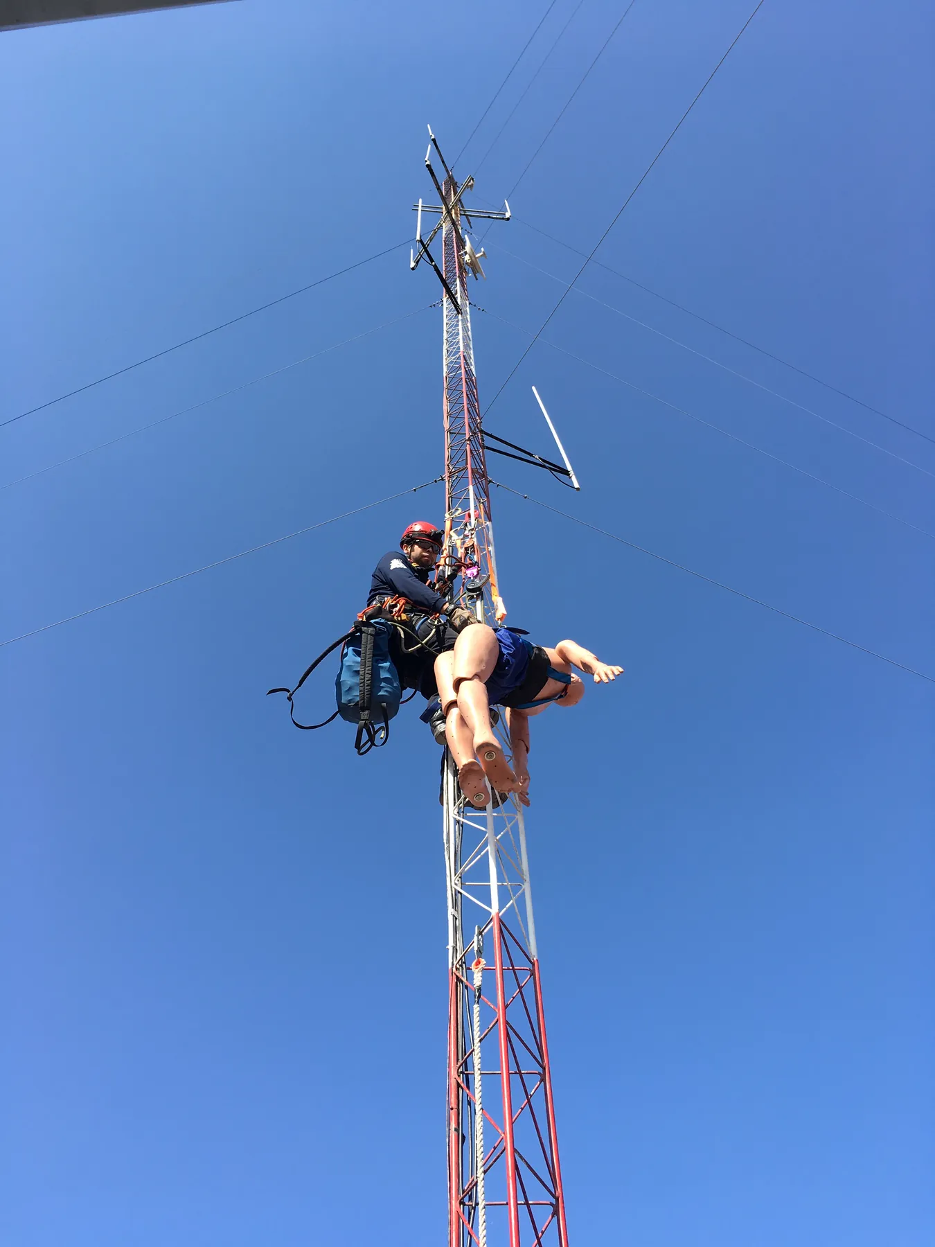 Rescatista suspendido en una torre de telecomunicaciones baja a un maniquí contra el cielo azul.