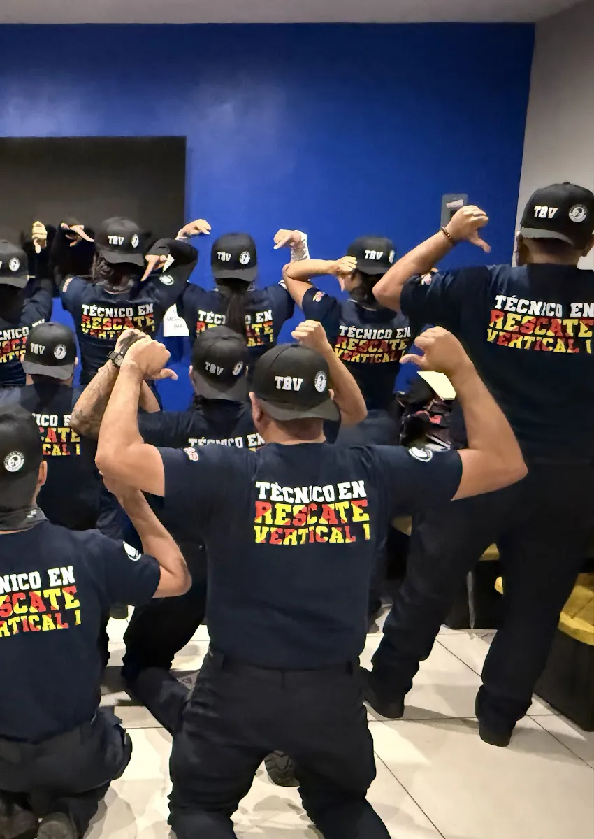 Alumnos de espaldas, con gorras «TRV» y playeras «Técnico en Rescate Vertical 1», se señalan la espalda frente a una pared azul.